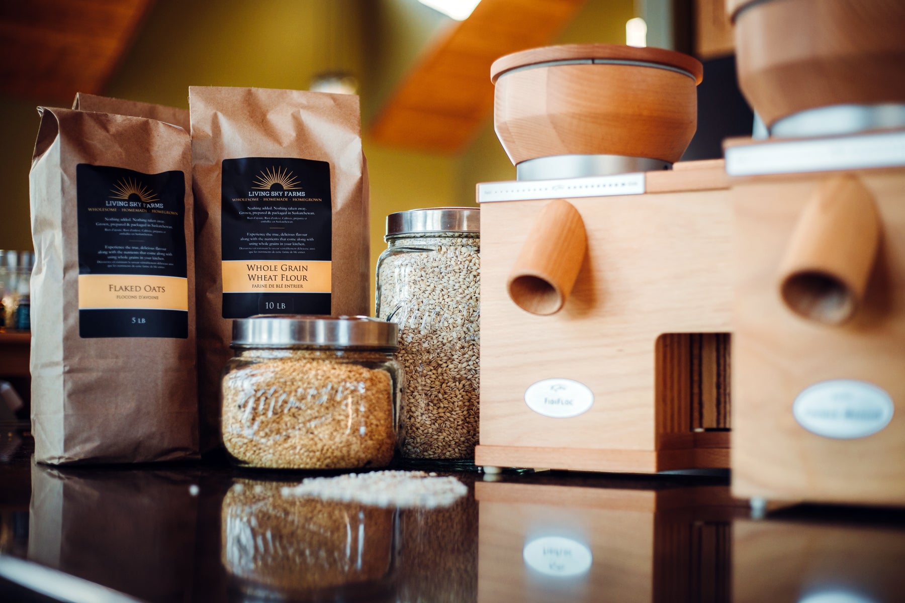 A package of flaked oats and whole grain wheat flour from Living Sky farms with a taller and shorter mason jar full of whole grains beside two Komo Mills