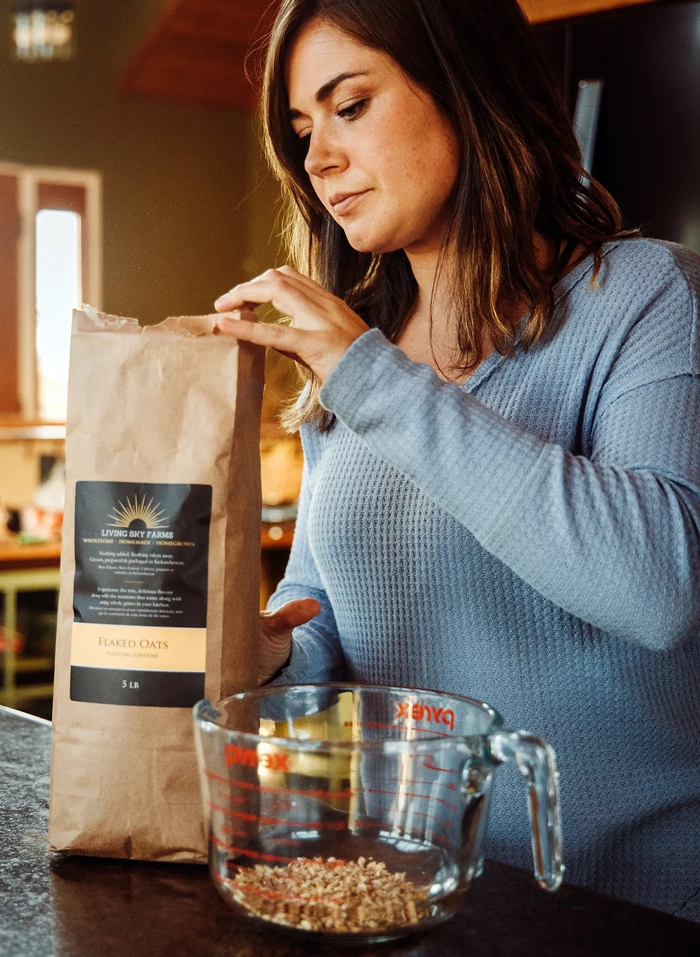 In a sunny kitchen, Sarah is wearing a blue sweater as she pours a bag of her flaked oats into a 5-cup measure. 
