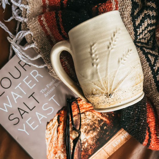 A harvest mug sitting on a red, beige and black blanket with a book called "Flour, water, salt, yeast" sitting next to it with a pair of black glasses on the book