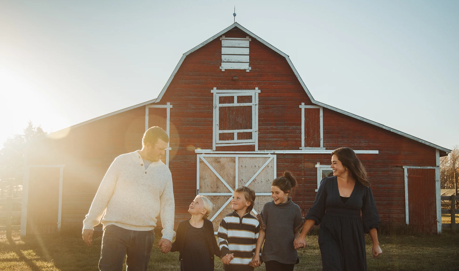 In the sunset, Sarah with her husband and three kids holding hands in front of their big red barn with white trim. 